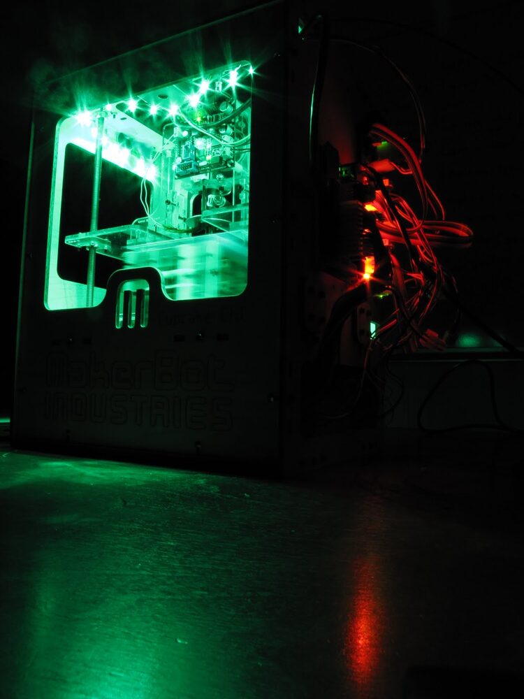 A MakerBot Cupcake CNC 3D printer lit with green LEDs, mid-print, photographed in a dark room around 2010.