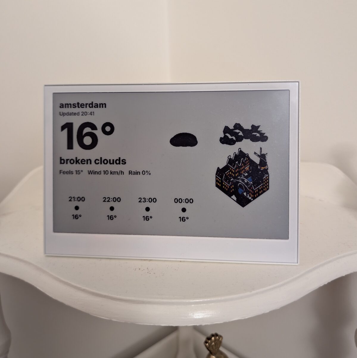 The reTerminal E1002 e-paper display on a side table, showing Amsterdam weather, an hourly forecast, and an AI-generated isometric illustration of canal houses.